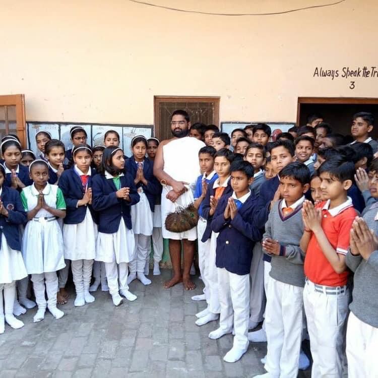 Maharaj Ji surrounded by schoolchildren in uniform with folded hands — school visit for value-based education on ahimsa and truth